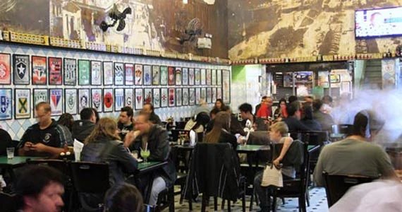 Salão movimentado de um bar, com clientes conversando em mesas e quadros decorativos nas paredes